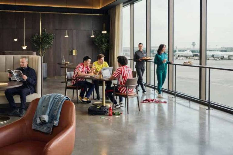 Group of travelers in a modern airport lounge around a table, some using laptops, with large windows and planes outside.