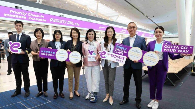 Eight people in business attire stand in a row at an airport, holding purple promotional signs for HK Express.",