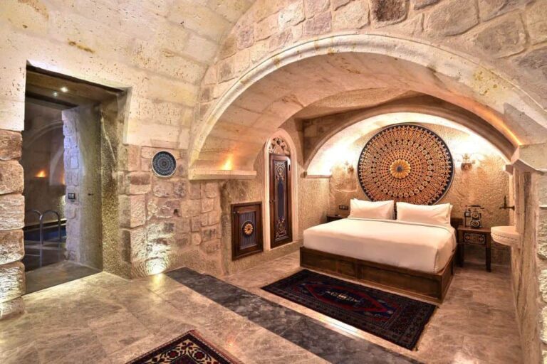 Stone-walled bedroom with a vaulted arch ceiling, king bed with white linens, and a large circular mandala headboard above it. Two nightstands, carved wooden doors, and Persian rugs complete the rustic, cave-like interior.] ,