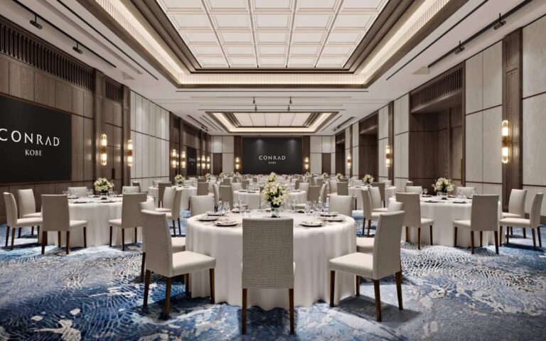 Elegant banquet hall with round tables covered in white cloth and floral centerpieces, Conrad Kobe backdrop in the distance.