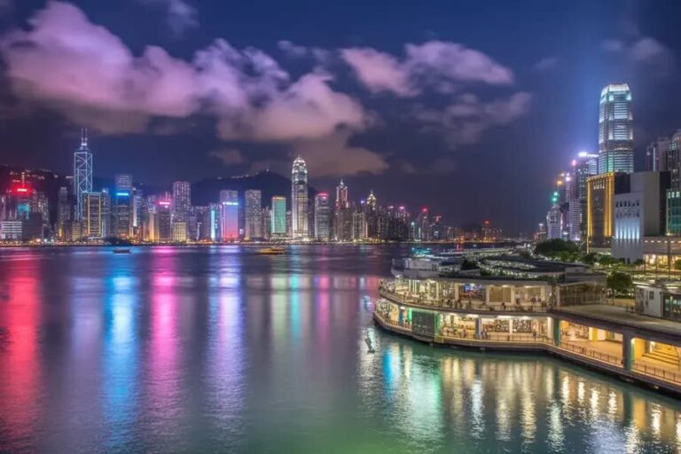 Night view of a vibrant city skyline along a harbor, with colorful reflections dancing on the water and a lit waterfront promenade.