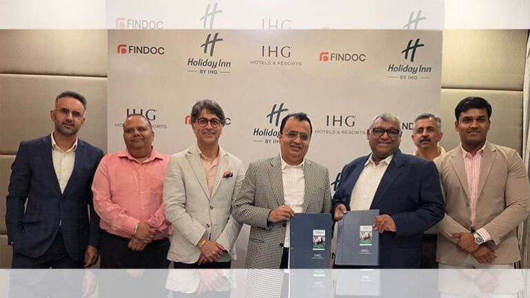 Seven men in business attire pose for a photo at a corporate event, standing in front of an IHG/Holiday Inn backdrop; two men hold blue folders.