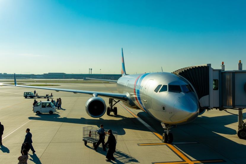Airline passenger jet parked at a gate with a jet bridge attached, ground crew and travelers nearby.