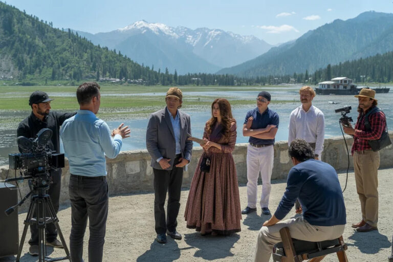 Film crew interviews two actors in period clothing beside a lake with mountains in the background.