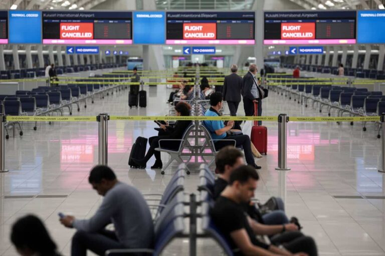 Airport terminal with rows of seats, yellow barrier tape, and passengers waiting as flight cancellation signs appear on large screens in the background.