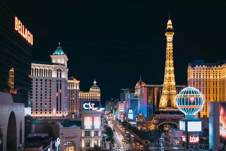 Nighttime Las Vegas street scene with the Eiffel Tower replica, Bellagio sign, and neon-lit casinos along a busy boulevard.