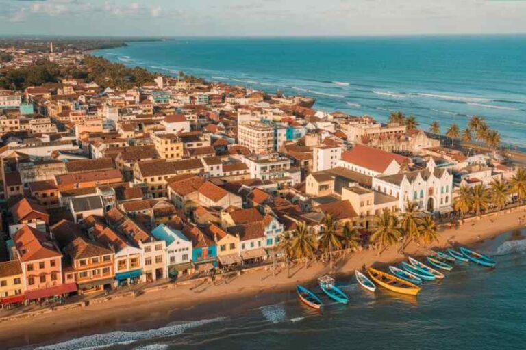 Aerial view of a seaside town with pastel-colored houses along a sandy beach and colorful boats docked at the shore.