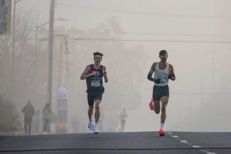 Two male runners race side by side on a misty road, bibs visible on their chests.