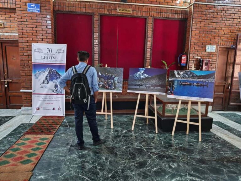 Man with backpack viewing four mounted landscape photos on easels in a brick-walled hall, with a tall banner on the left.