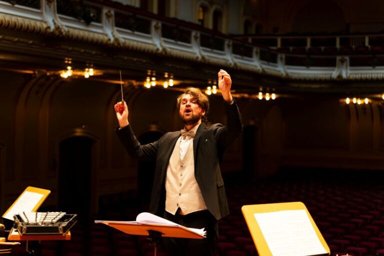 Conductor in a black suit and beige vest raises a baton while leading an orchestra on a warmly lit, ornate concert hall stage with sheet music stands nearby.