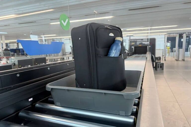 Black rolling suitcase standing upright in a gray bin on an airport baggage conveyor, with a water bottle in its side pocket.