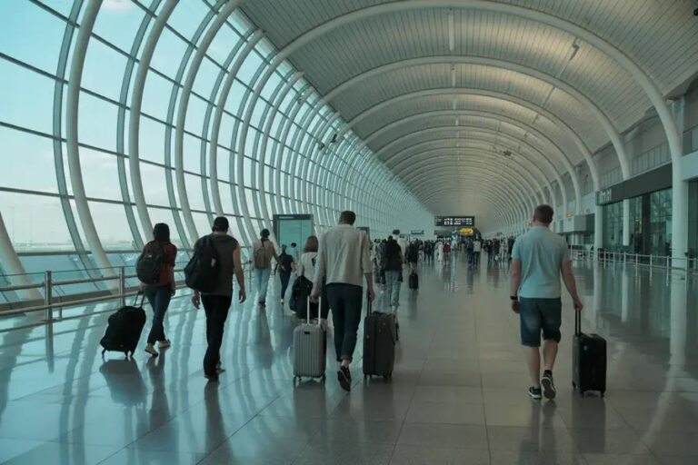 People walking with rolling suitcases through a spacious curved-airport terminal with glass walls at left.