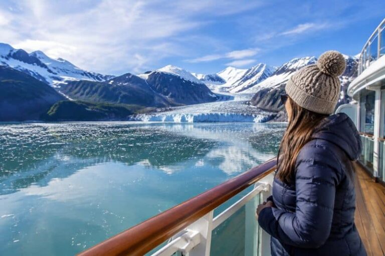 Woman in a blue puffer jacket and knit beanie on a cruise deck gazing at snowcapped mountains and a glacier-fed lake.