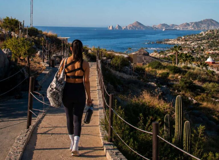 A woman in a black athletic outfit walks along a stone path toward the sea, carrying a large gym bag and water bottle, with a coastal town and blue ocean in the background.