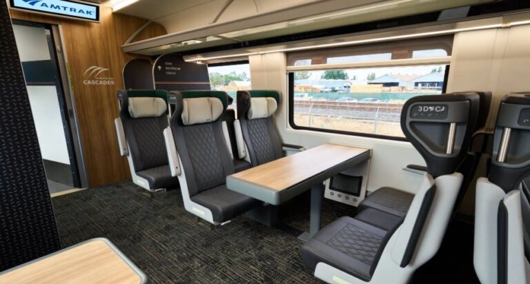 Interior of an Amtrak Cascades train car with row seats around a linked wooden table, window view of tracks outside, modern gray and beige seating.