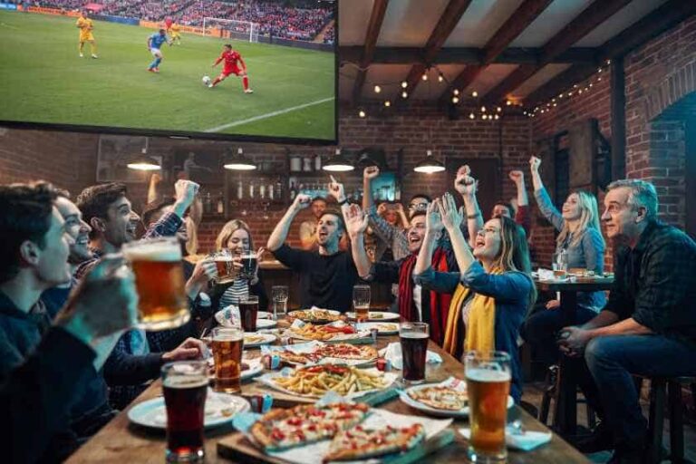 Group of friends in a sports bar cheering and raising their arms as a soccer match plays on a big screen, with pizza and drinks on the table.