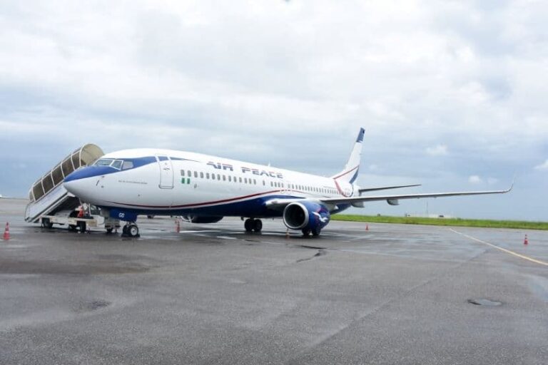 White passenger jet with mobile boarding stairs at the front door parked on a wet tarmac, under a cloudy sky (Air Peace livery).