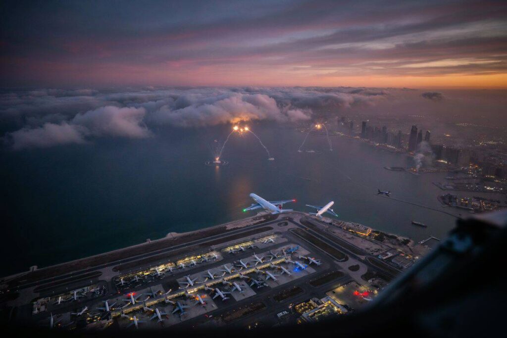Aerial view of an airport at sunset with multiple planes parked on the tarmac and a city skyline across the water in the distance