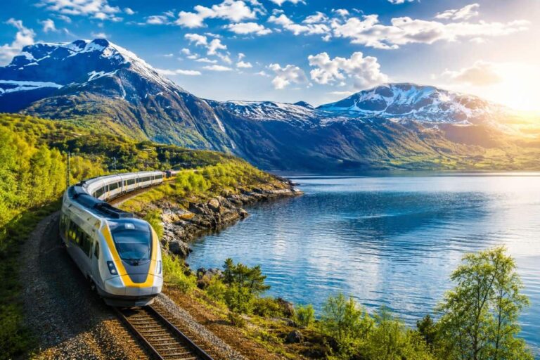 Modern white and yellow high-speed train curves along tracks beside a calm blue lake, with snow-capped mountains in the background under a blue sky and sun.