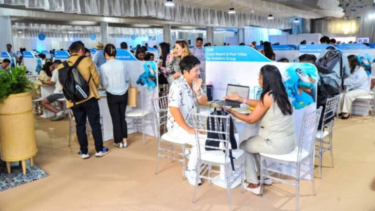 Crowded exhibition hall with travel booths; attendees talk to representatives at tables with laptops under blue signage.