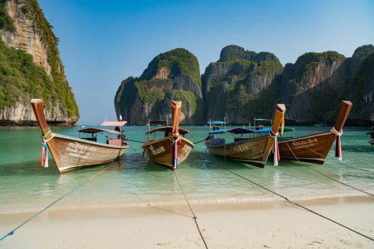 Four wooden long-tail boats tied to the shore on a tropical beach with turquoise water and towering limestone cliffs in the background with ribbons on the bows.