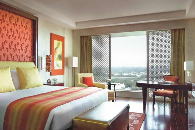 Hotel room with a king bed, orange/yellow accents, bench at foot, and a large window with patterned screens and city view.