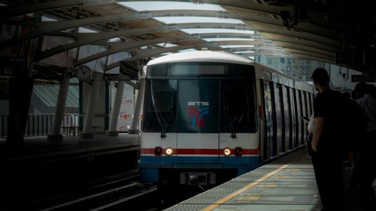 Train at an urban metro platform with BTS logo on the front and passengers waiting on the right side, early in the day light filtering through the station ceiling.