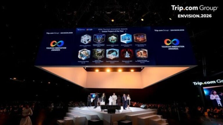 Stage at Trip.com Group ENVISION.2026 awards ceremony with presenters on a white platform and a large blue screen displaying award icons above the audience.