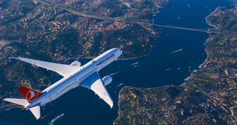 A Turkish Airlines passenger jet flying over a coastline and city from an aerial perspective.