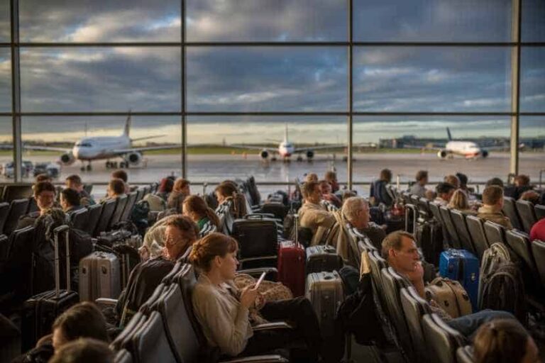 People seated in an airport terminal with rolling suitcases, waiting for boarding as planes sit on the runway outside the glass wall