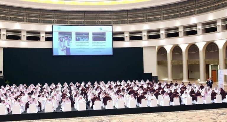 Conference hall filled with rows of attendees in white robes facing a stage with a large presentation screen above.