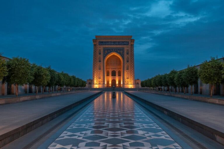 Night view of a tall, illuminated arch gateway at the end of a reflecting pool, flanked by tree-lined avenues. (Informative)
