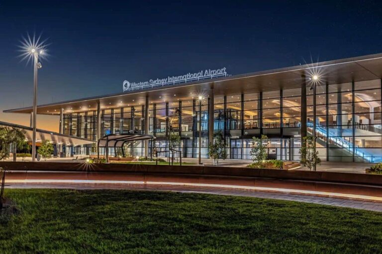 Night exterior of Western Sydney International Airport with glass-front terminal, bright lights, and signage visible above the entrance, trees and walkway in foreground