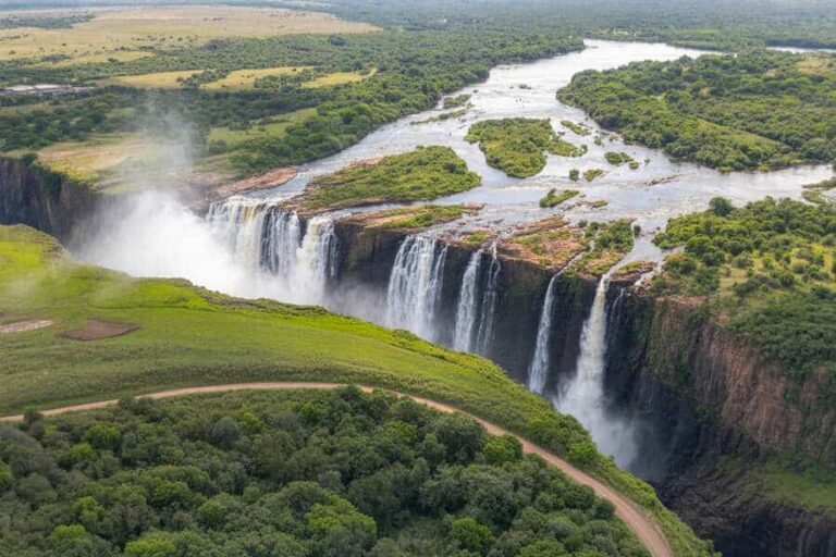 A wide waterfall spills over a cliff into a misty gorge, surrounded by lush green vegetation and a winding path below.