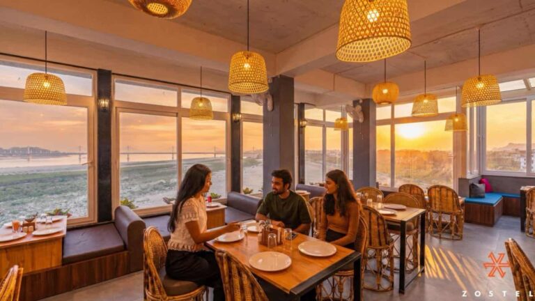 Three friends dining at a sunlit seaside restaurant with large windows and woven pendant lights during sunset.