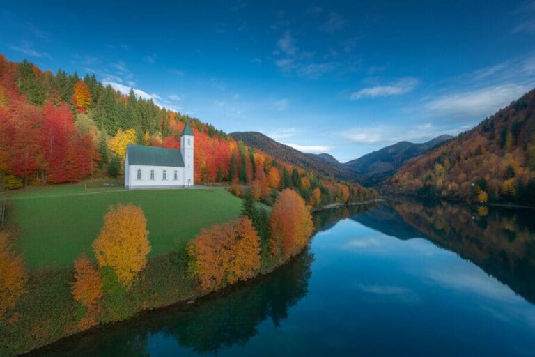 Autumn landscape with a white church on a grassy bank beside a calm lake, surrounded by colorful fall trees and mountains under a blue sky.