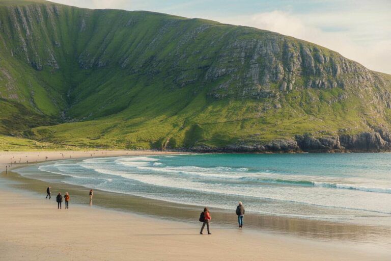 People strolling along a sandy beach with turquoise waves and a tall, green hillside cliff in the background.