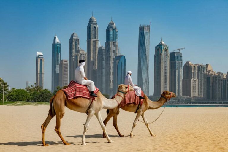 Two camels with riders walk along a sandy beach as a modern Dubai skyline rises in the background, under a blue sky