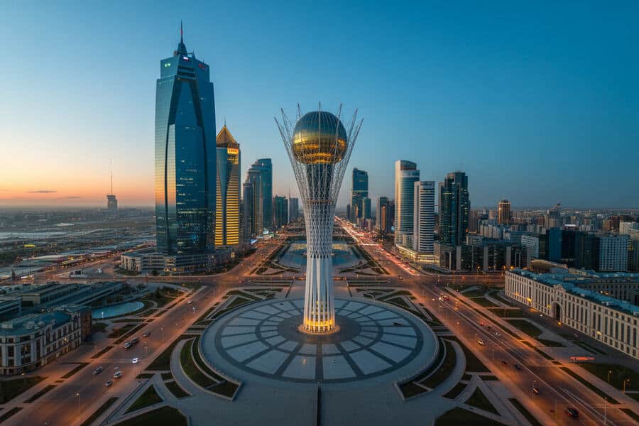 Aerial view of a modern city with a central Bayterek Tower surrounded by a circular plaza and tall skyscrapers at dusk in Astana, Kazakhstan.