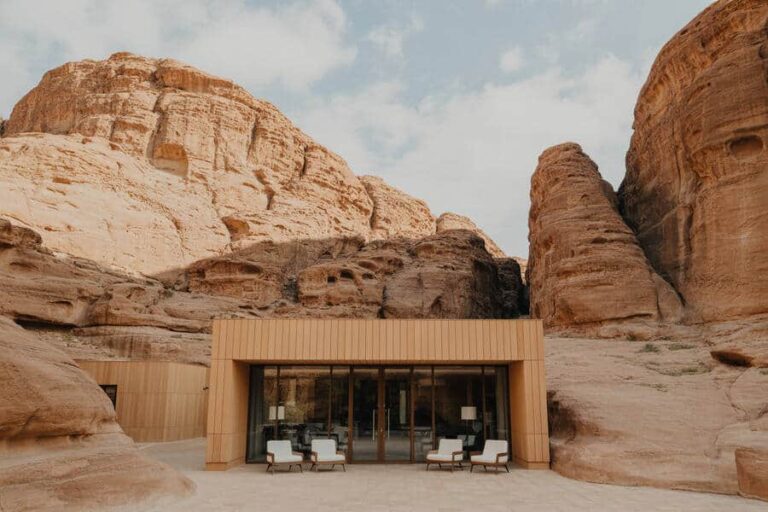 Modern rectangular glass-front house built into beige rock cliffs, with white outdoor lounge chairs outside.