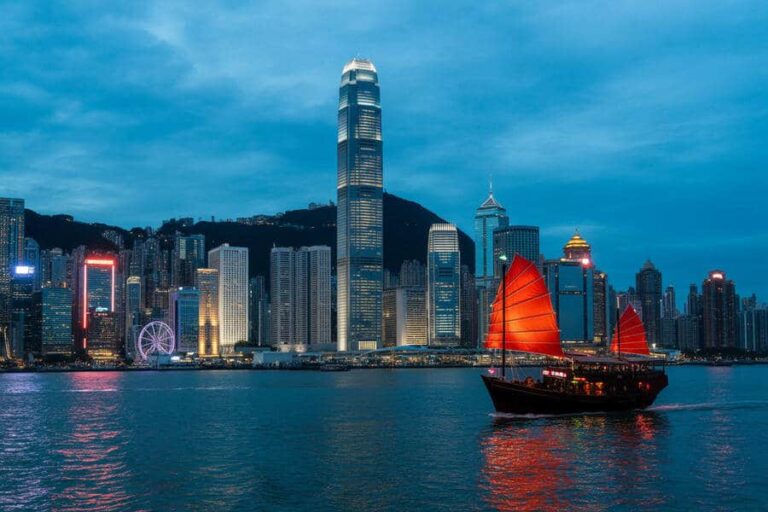 Hong Kong skyline at dusk with towering skyscrapers along Victoria Harbour and a red-sailed junk boat in the foreground.