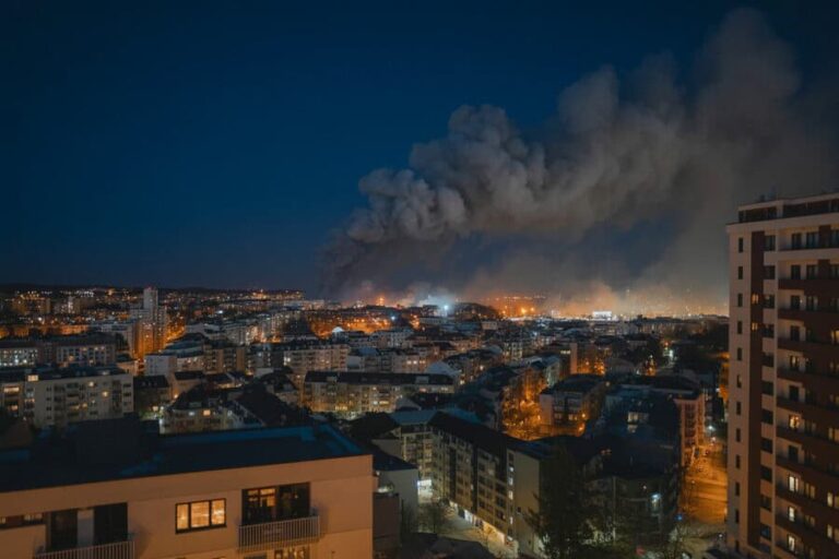 Nighttime cityscape with a large plume of smoke rising from a distant fire on the horizon.
