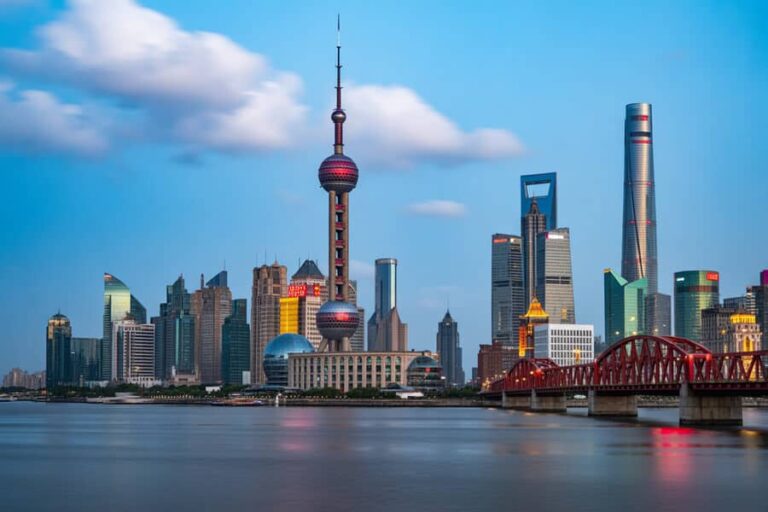 Shanghai skyline across the Huangpu River with the Oriental Pearl Tower and glass skyscrapers, a red bridge in the foreground.