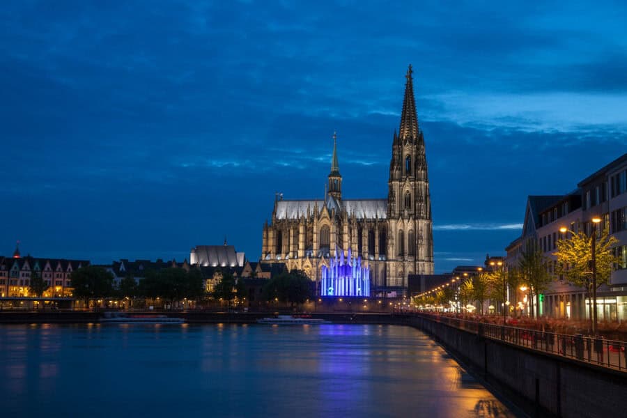 Gothic cathedral illuminated at dusk along a calm river, with a lined promenade and city lights.