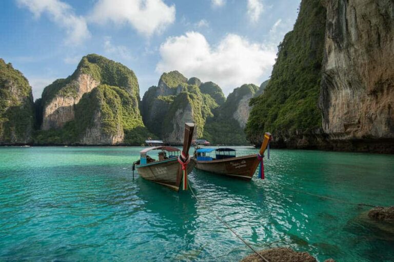 Two traditional Thai long-tail boats anchored in turquoise lagoon with towering green karst cliffs behind them.