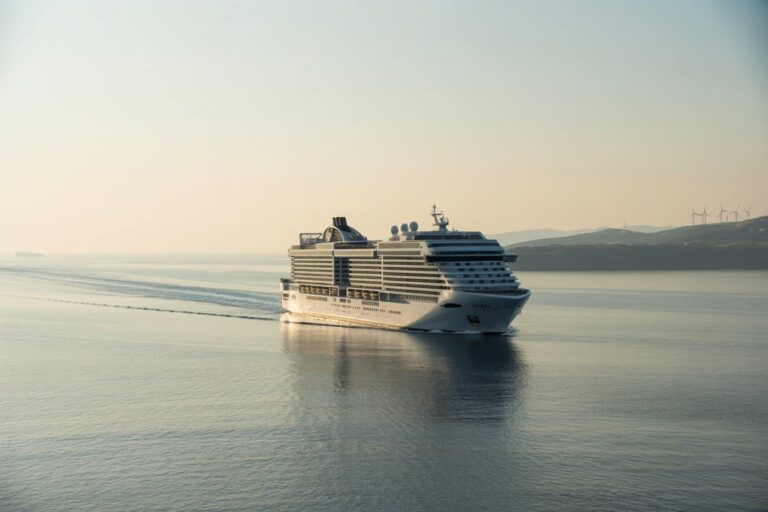 Large white cruise ship gliding on calm water with a distant coastline and wind turbines on the horizon.