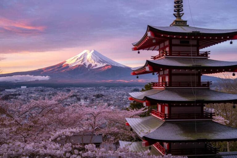 Snow-capped Mount Fuji rises over a city framed by a traditional red and white Japanese pagoda and blooming cherry trees at sunset.
