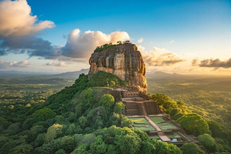 Rock fortress on a towering granite pillar rising from a dense jungle, with terraces and stairs ascending the slope at sunset.