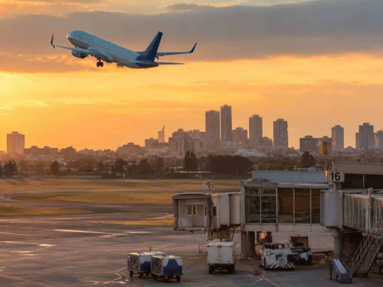 Airplane lifting off at sunset over a city skyline, with the airport jet bridge and tarmac in the foreground.