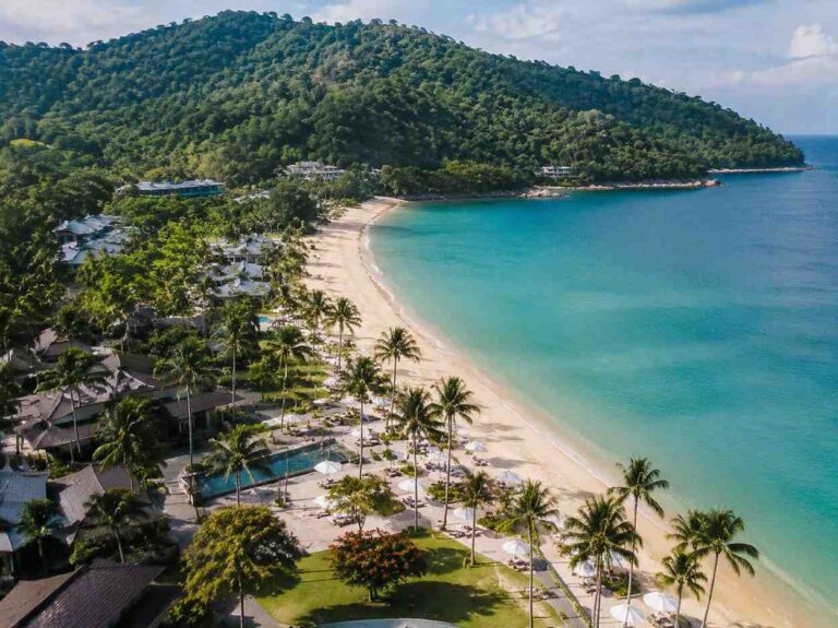 A tropical beach with turquoise water, white sand, and palm trees lining a beachside resort set against green hills.
